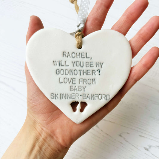 Personalised Godmother proposal gift, pearlised white clay hanging heart with baby feet cut out of the bottom, the heart is personalised with RACHEL, WILL YOU BE MY GODMOTHER? LOVE FROM BABY SKINNER-BAMFORD