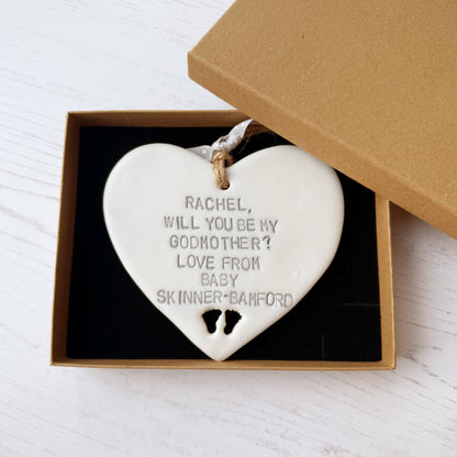 Personalised Godmother proposal gift, pearlised white clay hanging heart with baby feet cut out of the bottom, the heart is personalised with RACHEL, WILL YOU BE MY GODMOTHER? LOVE FROM BABY SKINNER-BAMFORD
