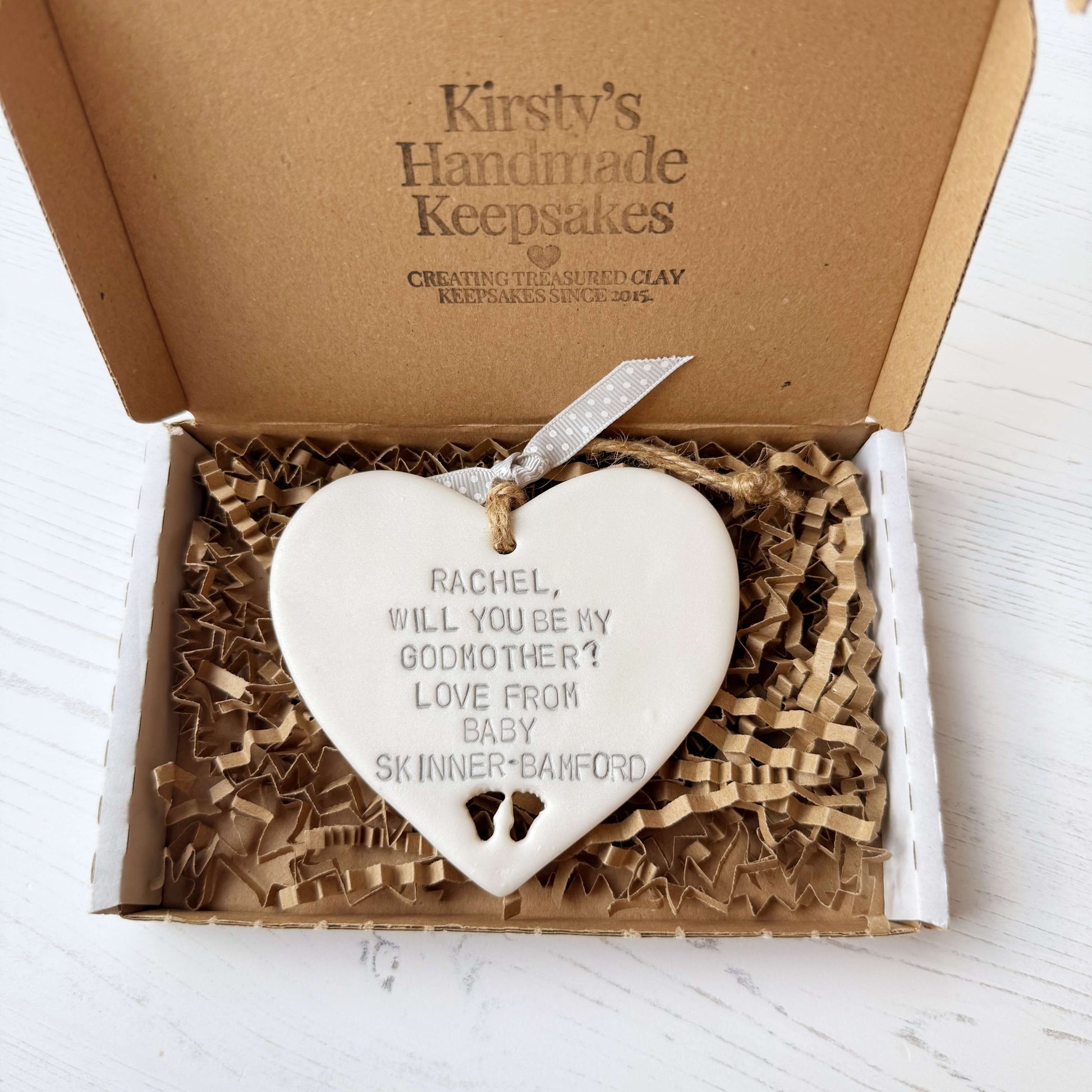 Personalised Godmother proposal gift, pearlised white clay hanging heart with baby feet cut out of the bottom, the heart is personalised with RACHEL, WILL YOU BE MY GODMOTHER? LOVE FROM BABY SKINNER-BAMFORD