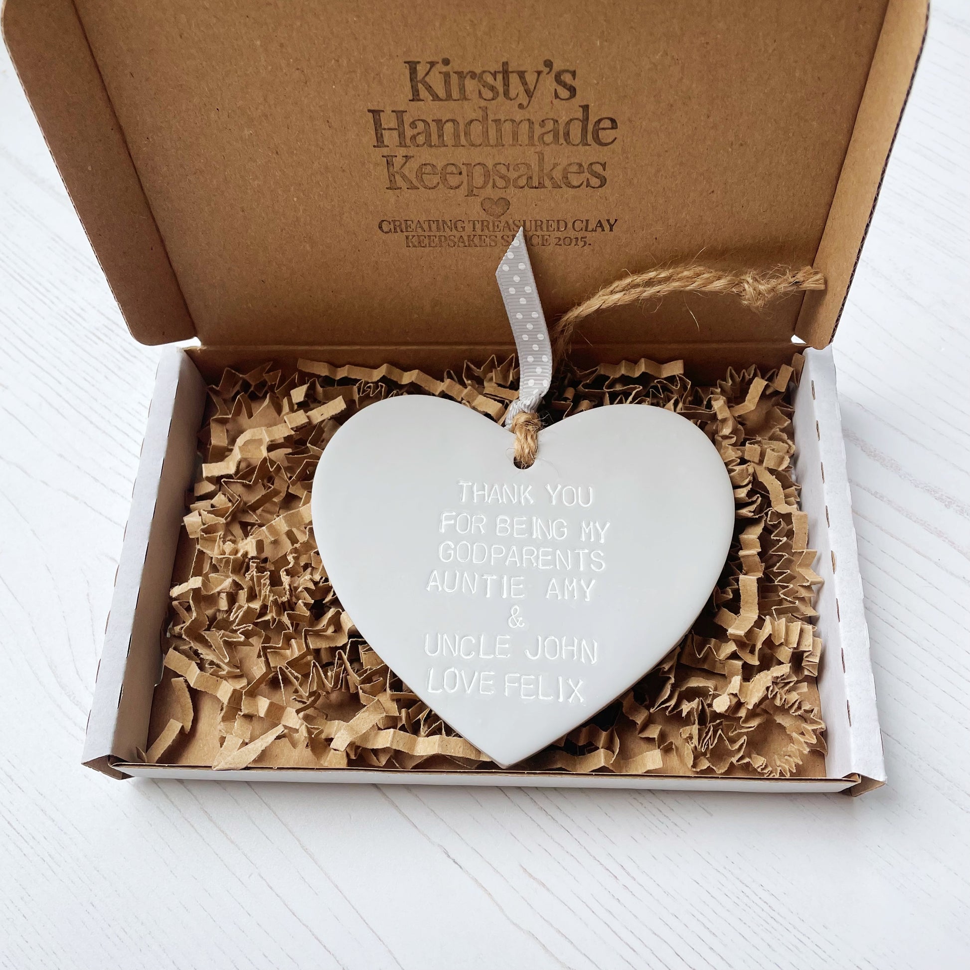 Personalised Godparents gift, grey clay hanging heart personalised in white with THANK YOU FOR BEING MY GODPARENTS AUNTIE AMY & UNCLE JOHN LOVE FELIX In a postal box with Kraft brown shredded zigzag paper 