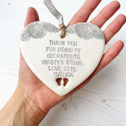 Personalised godparents gift, pearlised white clay hanging heart with baby feet cut out of the bottom and grey lace edge at the top of the heart, the heart is personalised with THANK YOU FOR BEING MY GODPARENTS KIRSTY & STEVE. LOVE OTTO 11/08/24