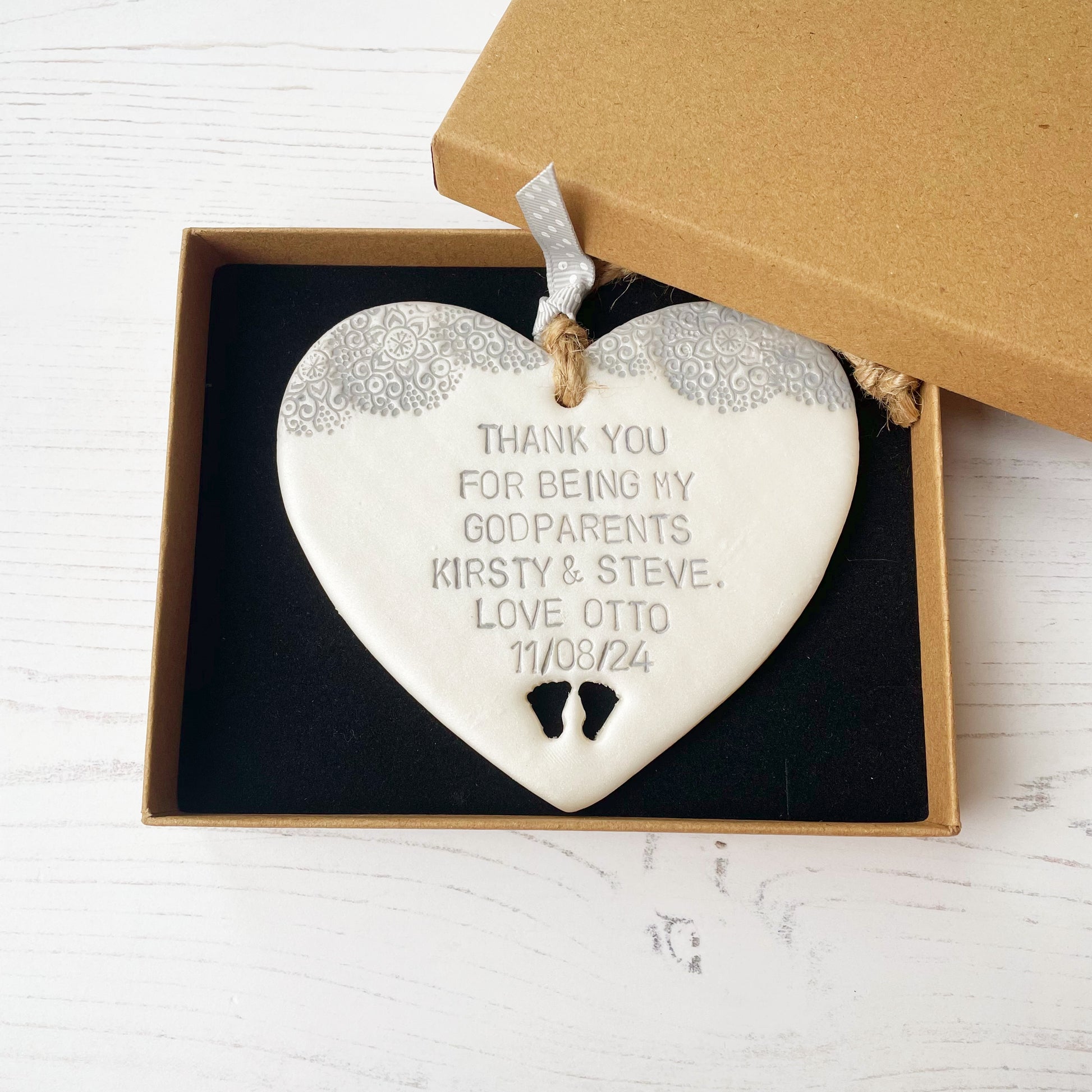 Personalised godparents gift, pearlised white clay hanging heart with baby feet cut out of the bottom and grey lace edge at the top of the heart, the heart is personalised with THANK YOU FOR BEING MY GODPARENTS KIRSTY & STEVE. LOVE OTTO 11/08/24