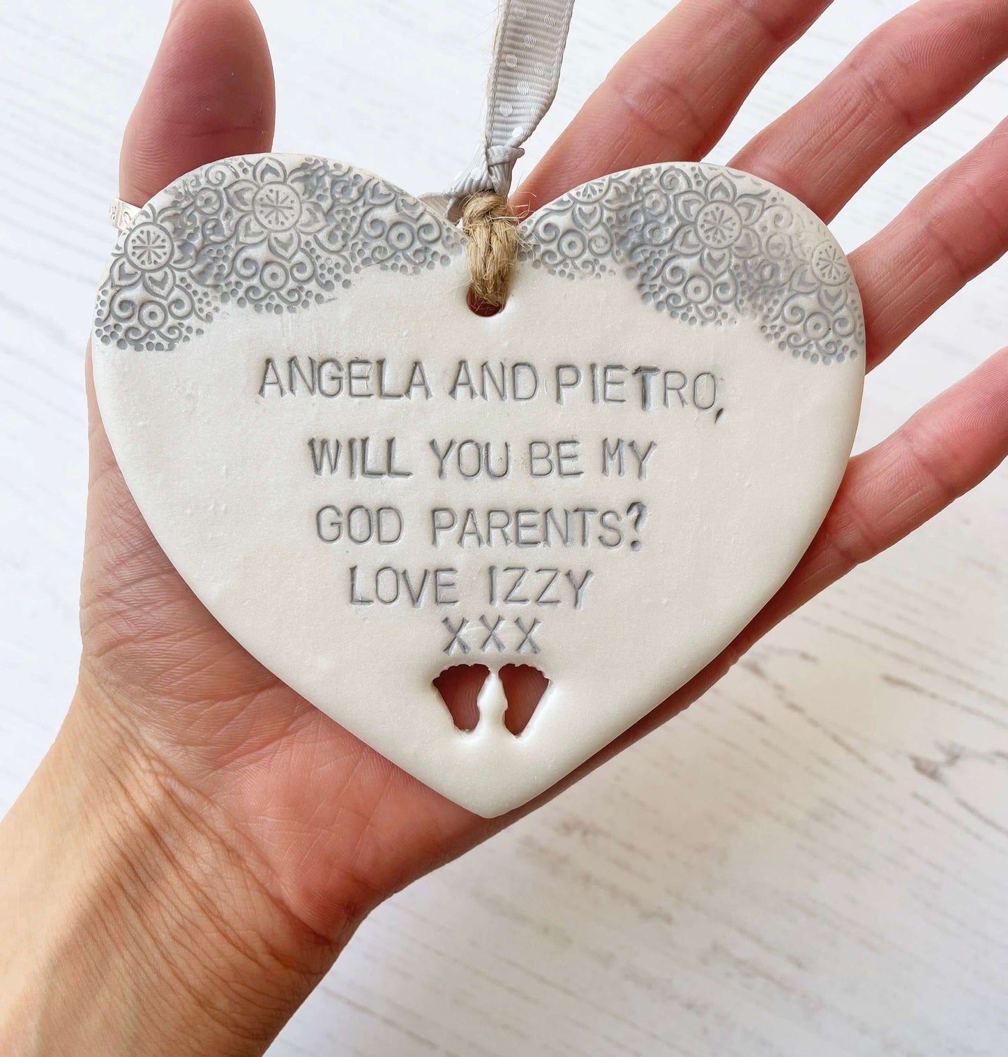 Will you be my godparents gift pearlised white clay hanging heart with baby feet cut out of the bottom grey lace edge at the top the heart is personalised with ANGELA & PIETRO, WILL YOU BE MY GODPARENTS? LOVE IZZY XXX