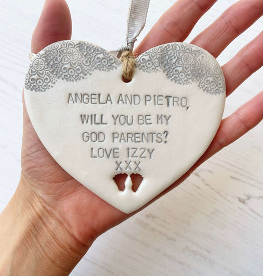 Will you be my godparents gift pearlised white clay hanging heart with baby feet cut out of the bottom grey lace edge at the top the heart is personalised with ANGELA & PIETRO, WILL YOU BE MY GODPARENTS? LOVE IZZY XXX