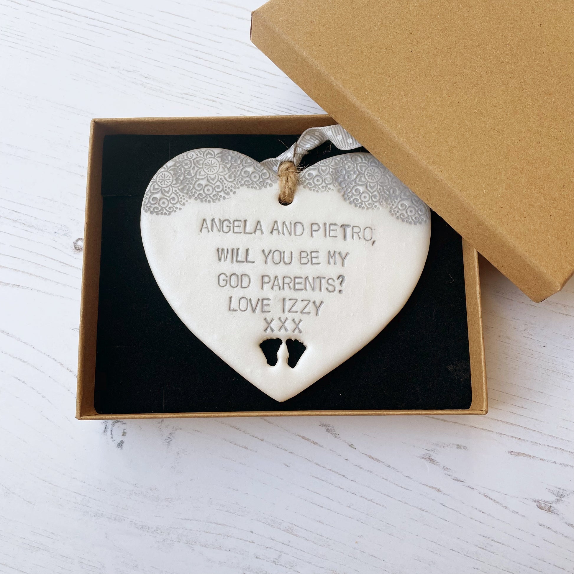 Will you be my godparents gift pearlised white clay hanging heart with baby feet cut out of the bottom grey lace edge at the top the heart is personalised with ANGELA & PIETRO, WILL YOU BE MY GODPARENTS? LOVE IZZY XXX