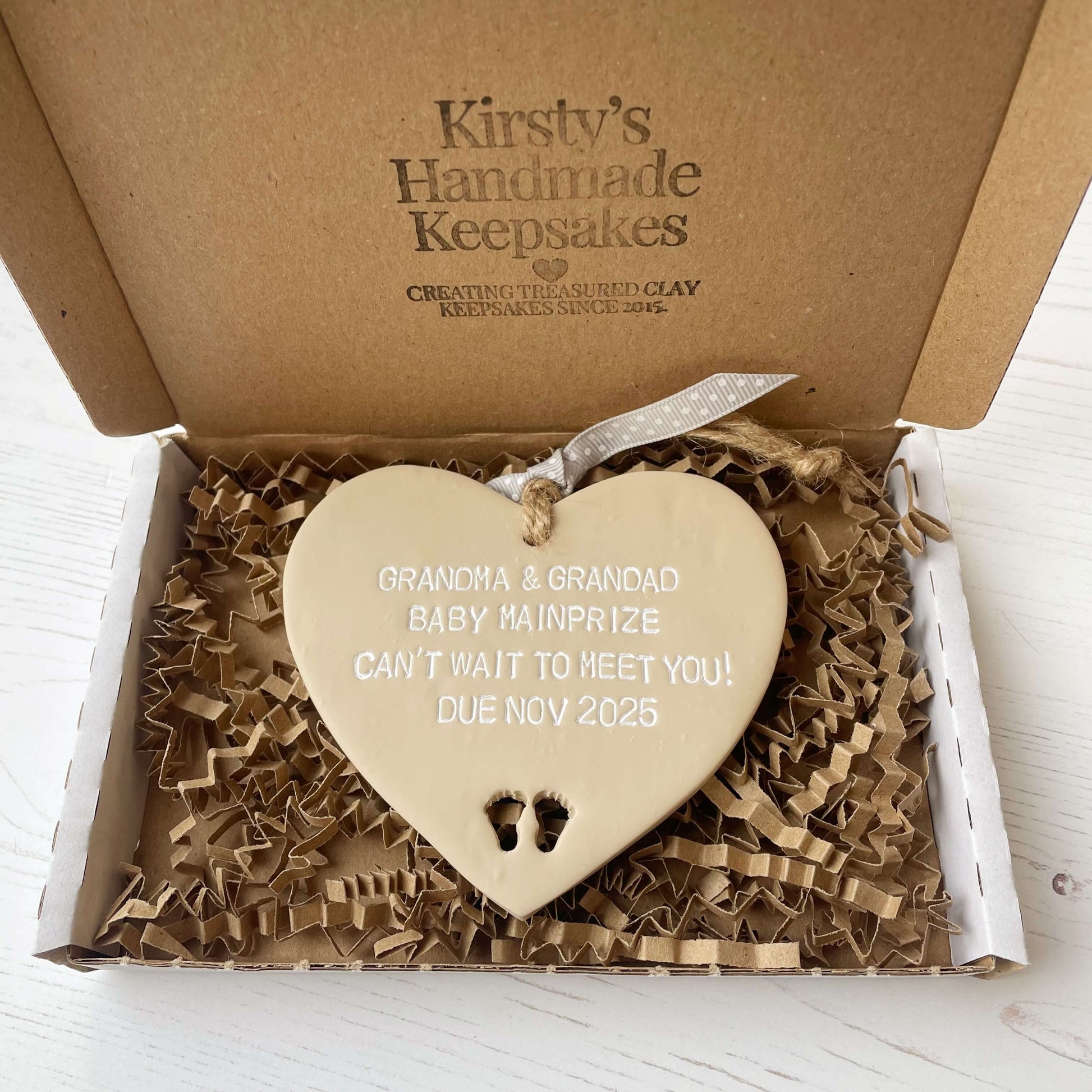 Beige clay hanging heart with baby feet cut out of the bottom and white personalisation, the heart is personalised with GRANDMA AND GRANDAD BABY MAINPRIZE CAN’T WAIT TO MEET YOU DUE NOV 2025