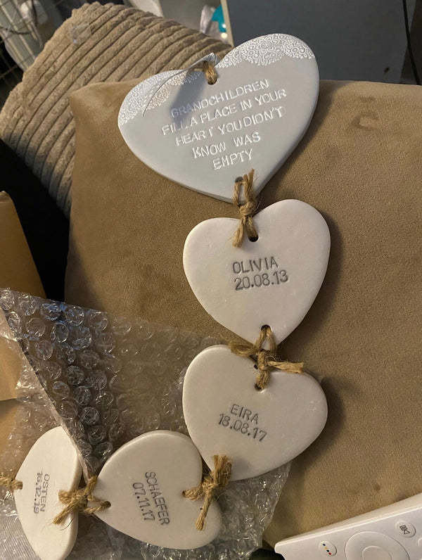 A set of personalised white polymer clay hearts hanging on a tan fabric surface, with one plaque still attached to bubble wrap packaging. The plaques are tied with twine and engraved with names, dates, and messages.
