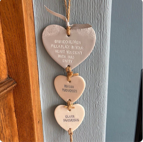 Three hanging polymer clay hearts  on a blue-grey wall next to a wooden doorframe. The top plaque is blank, while the middle and bottom plaques are personalised with engraved names and dates: 'Isaac 11/02/2023' and 'Clark 04/03/2025'.