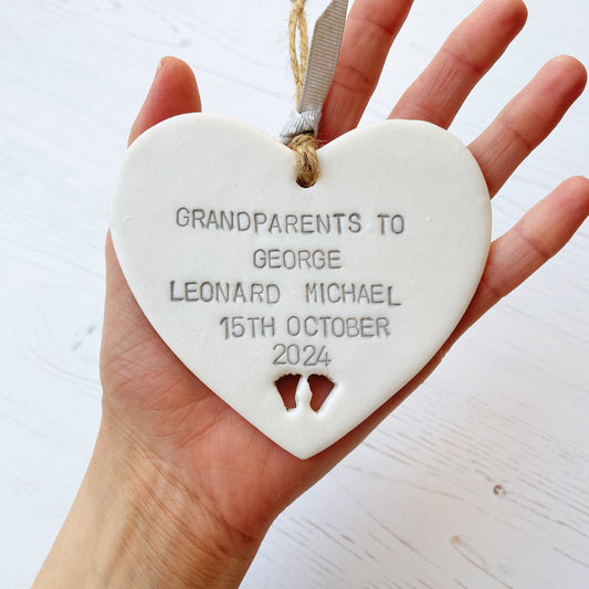 Pearlised white clay hanging heart with baby feet cut out of the bottom and grey personalisation, the heart is personalised with GRANDPARENTS TO GEORGE LEONARD MICHAEL 15TH OCTOBER 2024