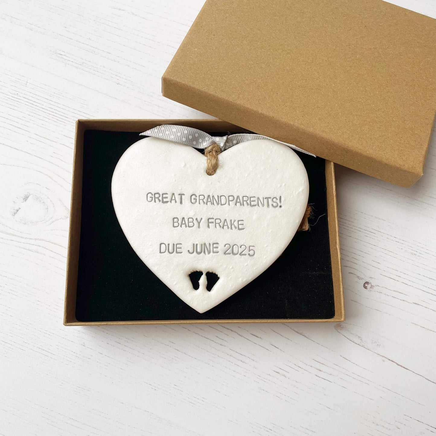 Pearlised white clay hanging heart with baby feet cut out of the bottom and grey personalisation, the heart is personalised with GREAT GRANDPARENTS! BABY FRAKE DUE JUNE 2025