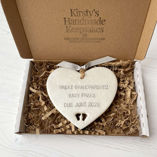 Pearlised white clay hanging heart with baby feet cut out of the bottom and grey personalisation, the heart is personalised with GREAT GRANDPARENTS! BABY FRAKE DUE JUNE 2025