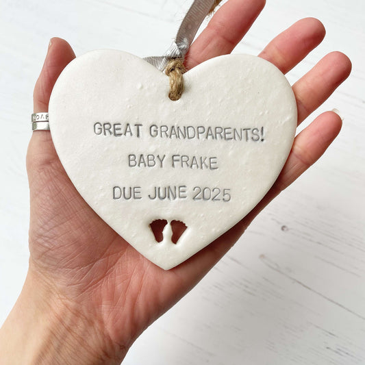 Pearlised white clay hanging heart with baby feet cut out of the bottom and grey personalisation, the heart is personalised with GREAT GRANDPARENTS! BABY FRAKE DUE JUNE 2025