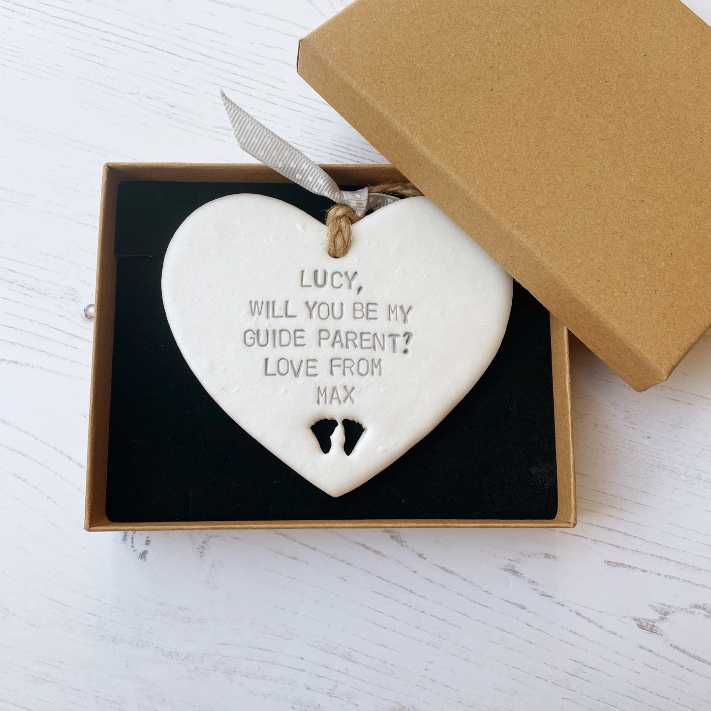 Personalised guide parent proposal gift, pearlised white clay hanging heart with baby feet cut out of the bottom, the heart is personalised with LUCY, WILL YOU BE MY GUIDE PARENT? LOVE FROM MAX