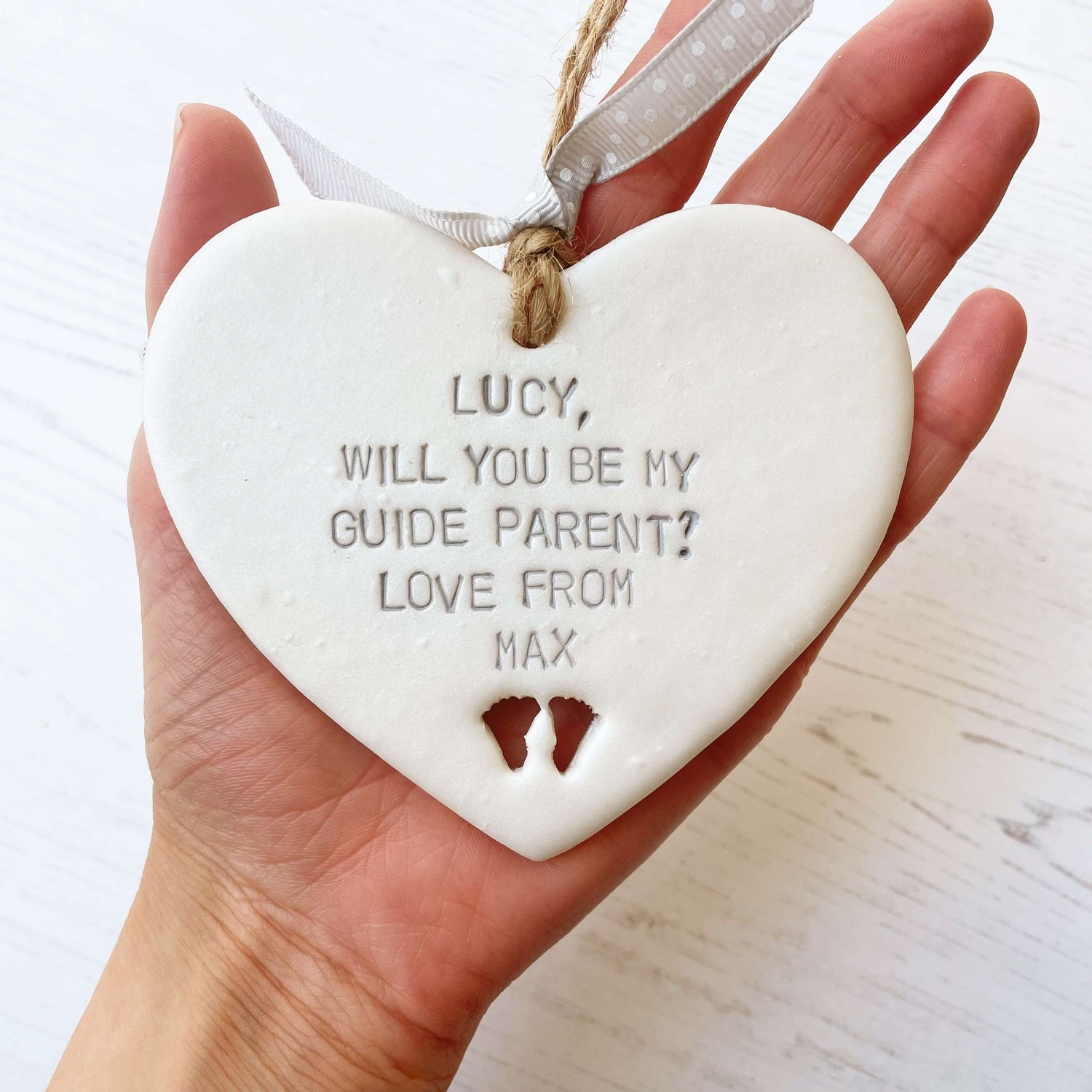 Personalised guide parent proposal gift, pearlised white clay hanging heart with baby feet cut out of the bottom, the heart is personalised with LUCY, WILL YOU BE MY GUIDE PARENT? LOVE FROM MAX