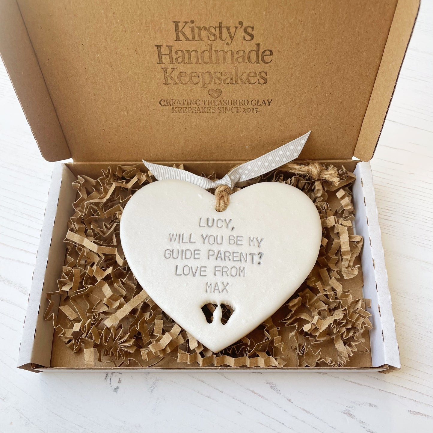 Personalised guide parent proposal gift, pearlised white clay hanging heart with baby feet cut out of the bottom, the heart is personalised with LUCY, WILL YOU BE MY GUIDE PARENT? LOVE FROM MAX