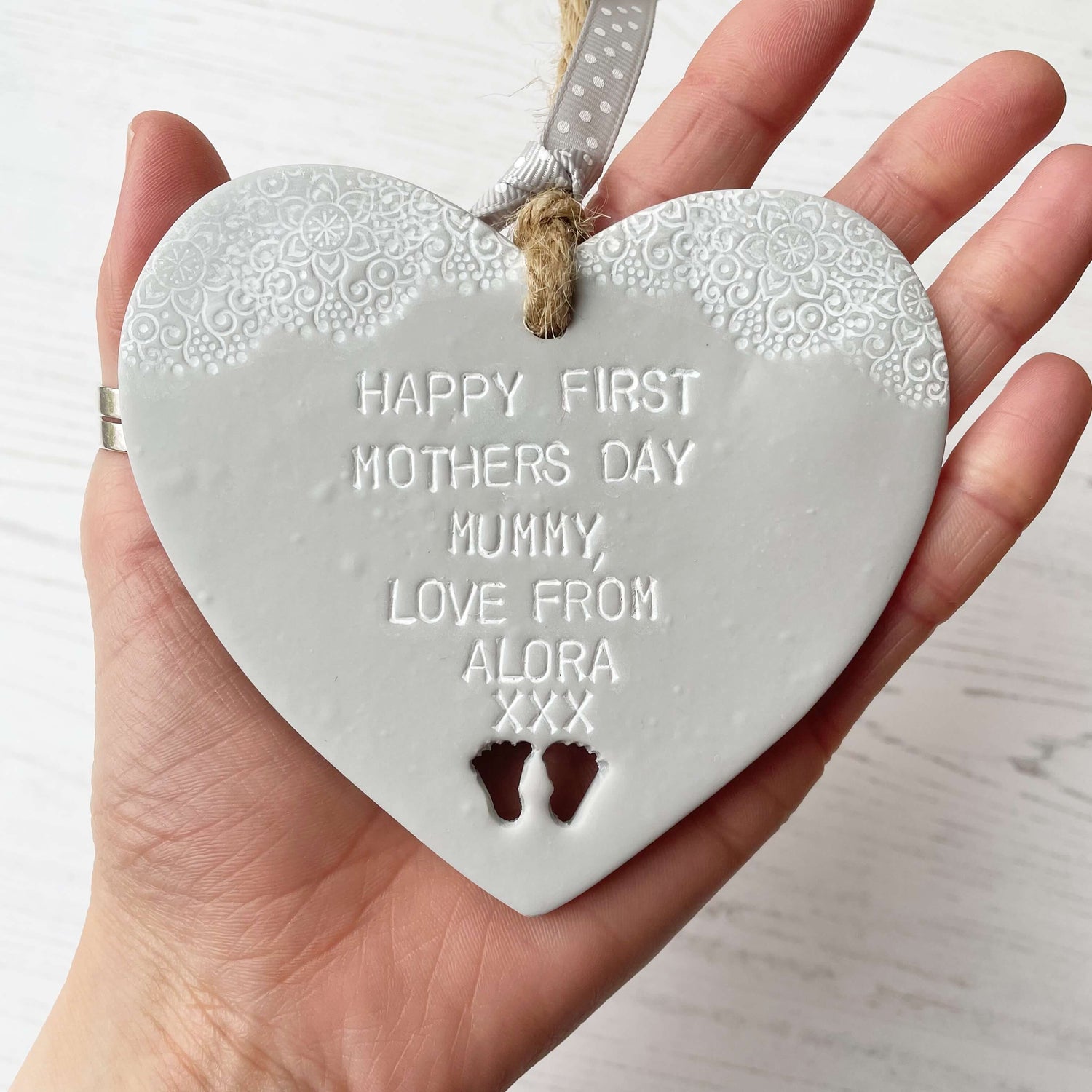 Personalised first Mother’s Day gift, grey clay hanging heart with baby feet cut out at the bottom of the heart and a white lace edge at the top of the heart, the heart is personalised with HAPPY FIRST MOTHERS DAY MUMMY, LOVE FROM ALORA XXX