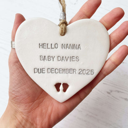 Pearlised white clay hanging heart with baby feet cut out of the bottom and grey personalisation, the heart is personalised with HELLO NANNA BABY DAVIES DUE DECEMBER 2025