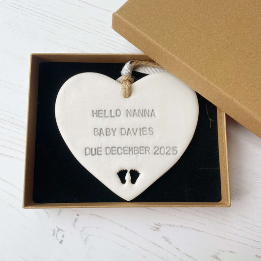 Pearlised white clay hanging heart with baby feet cut out of the bottom and grey personalisation, the heart is personalised with HELLO NANNA BABY DAVIES DUE DECEMBER 2025