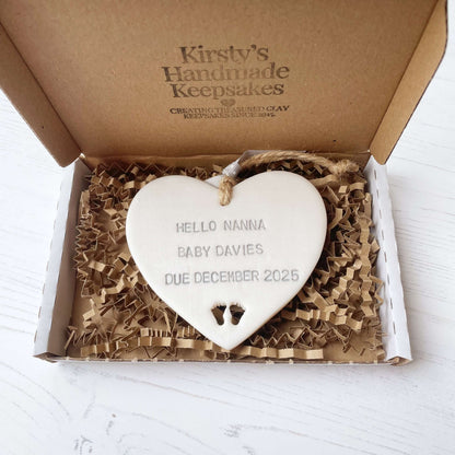 Pearlised white clay hanging heart with baby feet cut out of the bottom and grey personalisation, the heart is personalised with HELLO NANNA BABY DAVIES DUE DECEMBER 2025