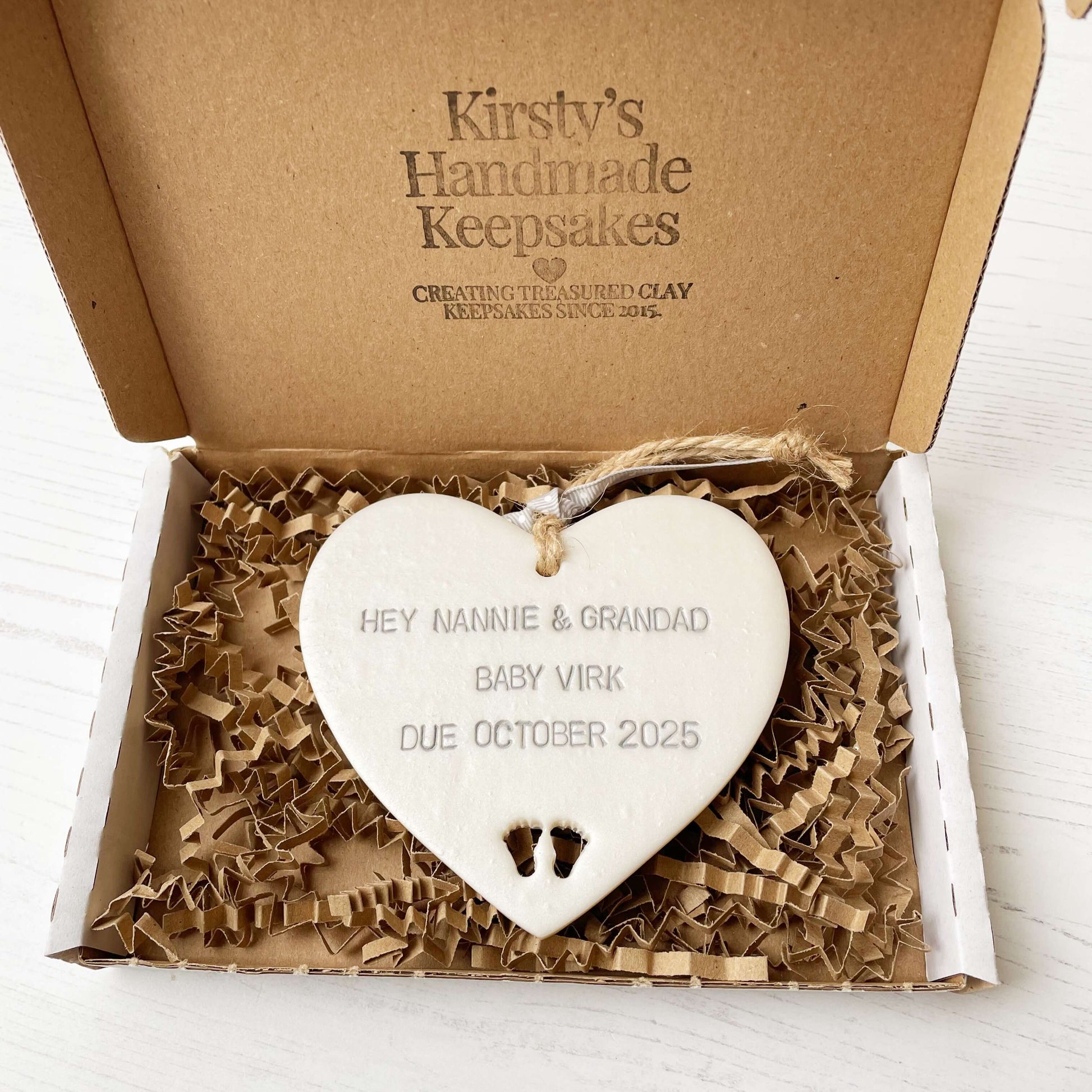 Pearlised white clay hanging heart with baby feet cut out of the bottom and grey personalisation, the heart is personalised with HEY NANNIE & GRANDAD BABY VIRK DUE OCTOBER 2025