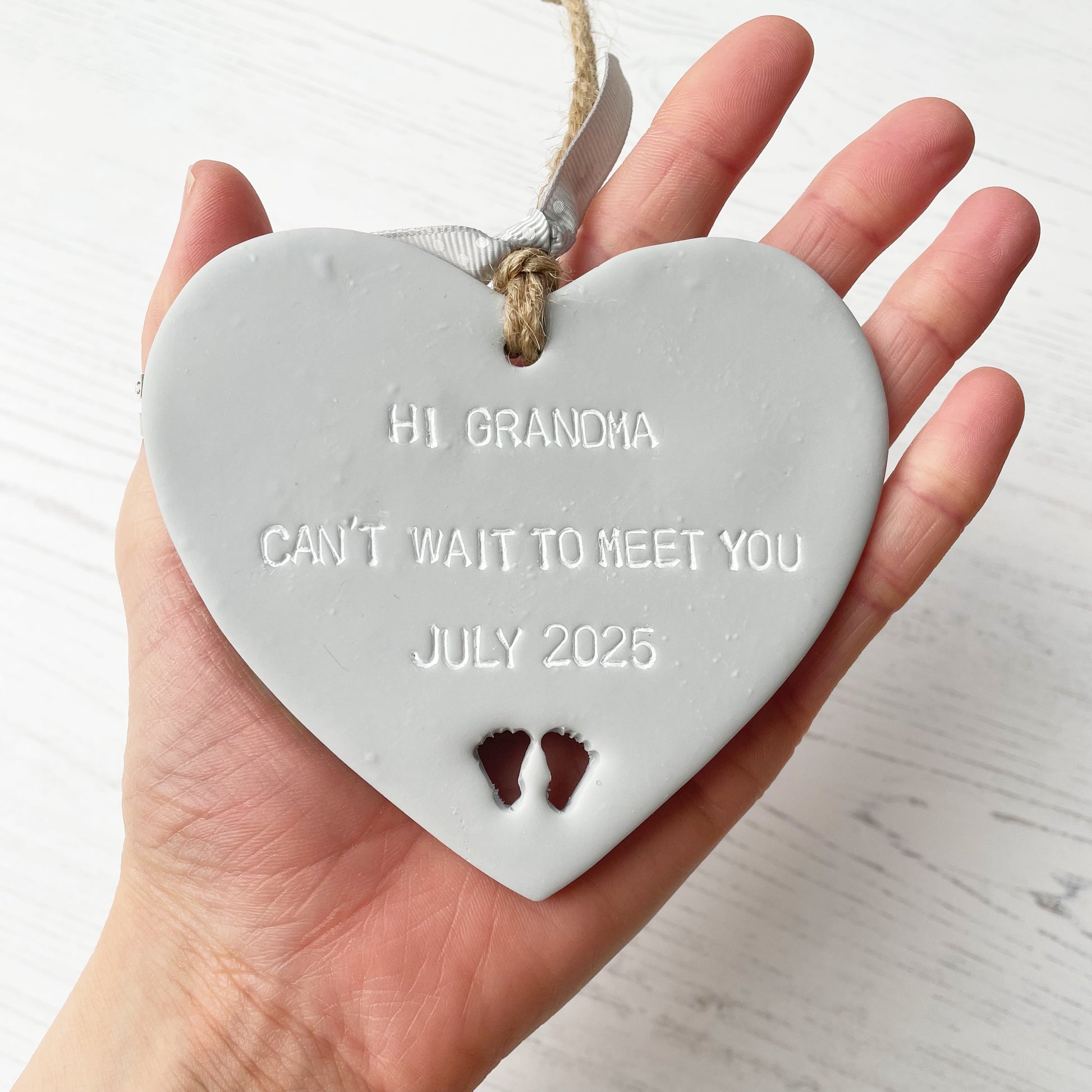 Grey clay hanging heart with baby feet cut out of the bottom and white personalisation, the heart is personalised with HI GRANDMA CAN’T WAIT TO MEET YOU JULY 2025