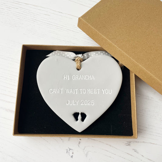 Grey clay hanging heart with baby feet cut out of the bottom and white personalisation, the heart is personalised with HI GRANDMA CAN’T WAIT TO MEET YOU JULY 2025