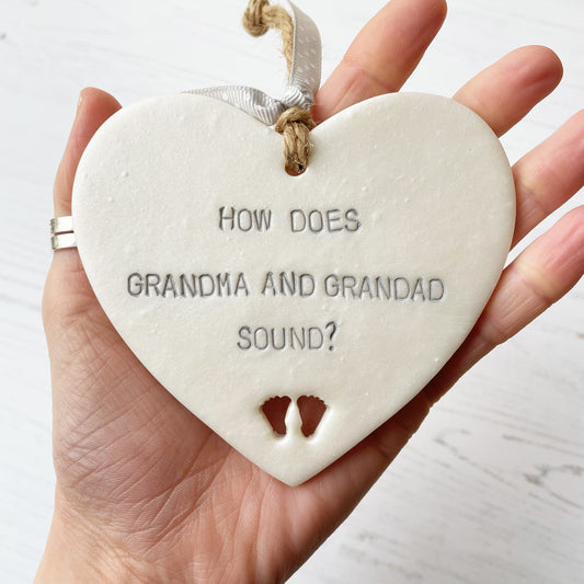 Pearlised white clay hanging heart with baby feet cut out of the bottom and grey personalisation, the heart is personalised with HOW DOES GRANDMA AND GRANDAD SOUND?