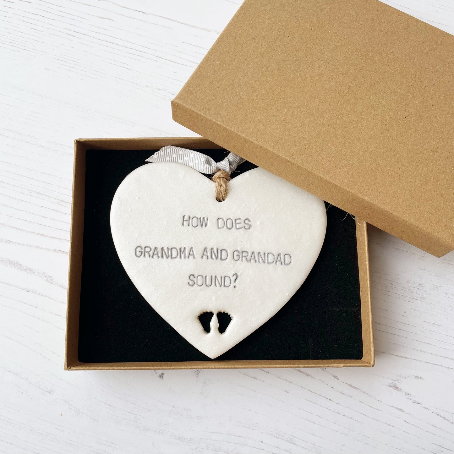 Pearlised white clay hanging heart with baby feet cut out of the bottom and grey personalisation, the heart is personalised with HOW DOES GRANDMA AND GRANDAD SOUND?