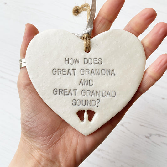 Pearlised white clay hanging heart with baby feet cut out of the bottom and grey personalisation, the heart is personalised with HOW DOES GREAT GRANDMA AND GREAT GRANDAD SOUND?