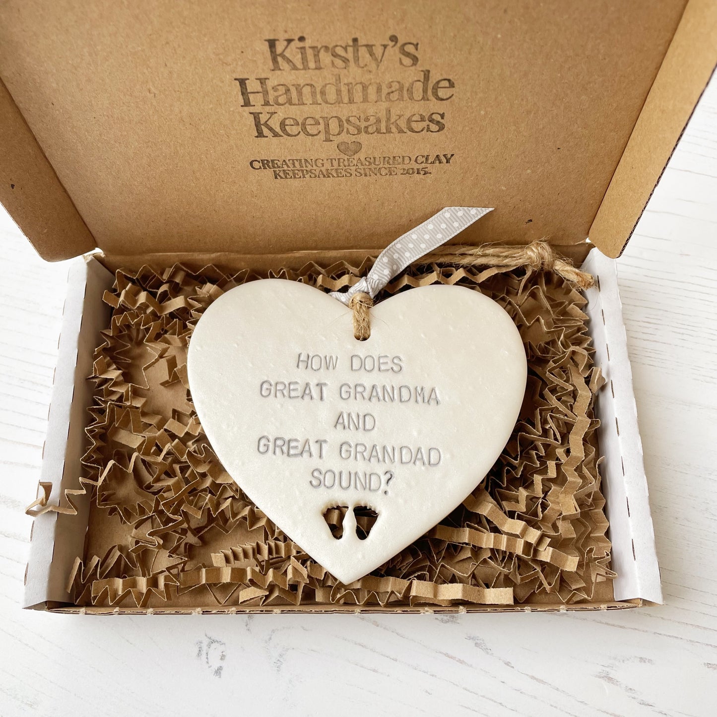 Pearlised white clay hanging heart with baby feet cut out of the bottom and grey personalisation, the heart is personalised with HOW DOES GREAT GRANDMA AND GREAT GRANDAD SOUND?