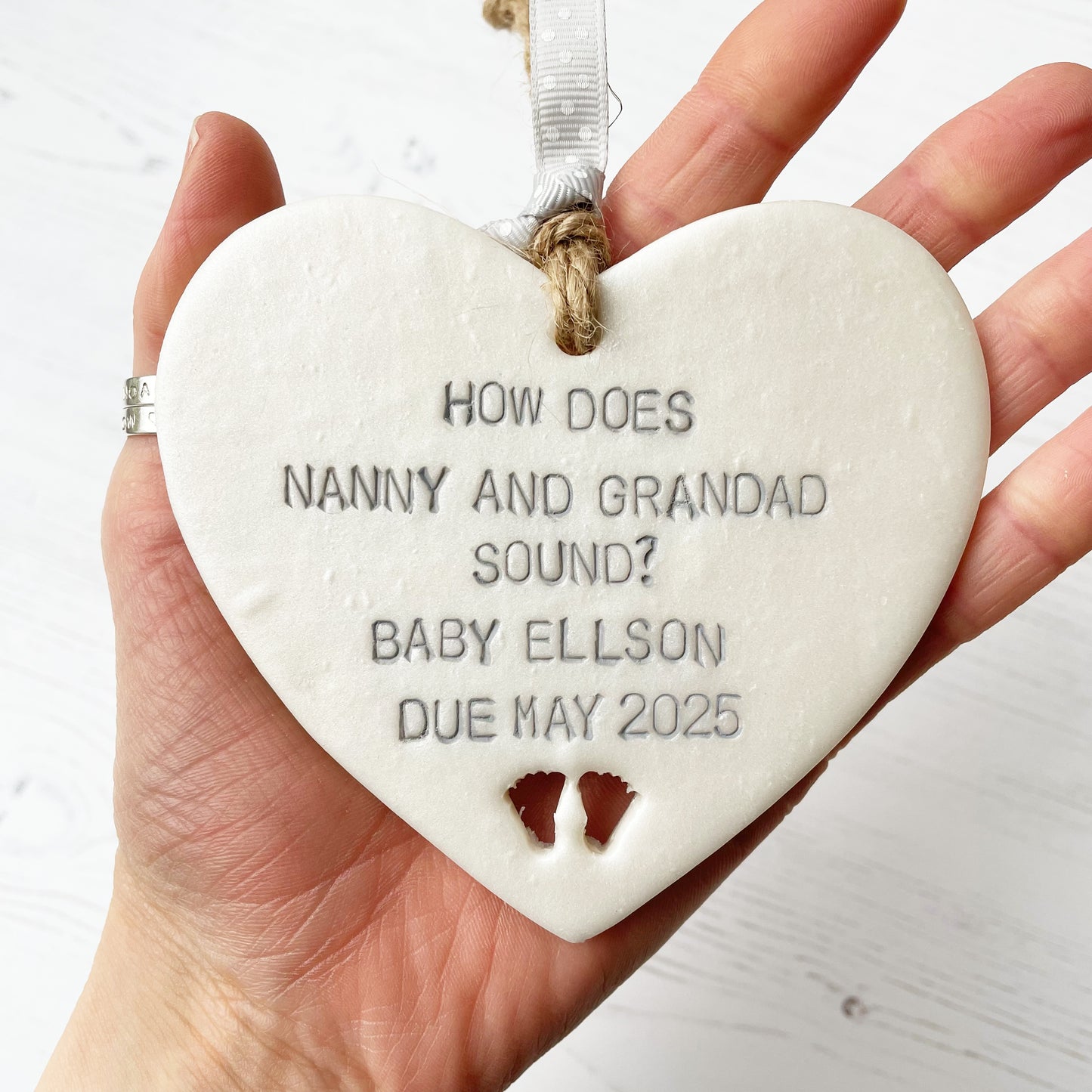 Pearlised white clay hanging heart with baby feet cut out of the bottom and grey personalisation, the heart is personalised with HOW DOES NANNY & GRANDAD SOUND? BABY ELLSON DUE MAY 2025