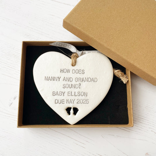 Pearlised white clay hanging heart with baby feet cut out of the bottom and grey personalisation, the heart is personalised with HOW DOES NANNY & GRANDAD SOUND? BABY ELLSON DUE MAY 2025