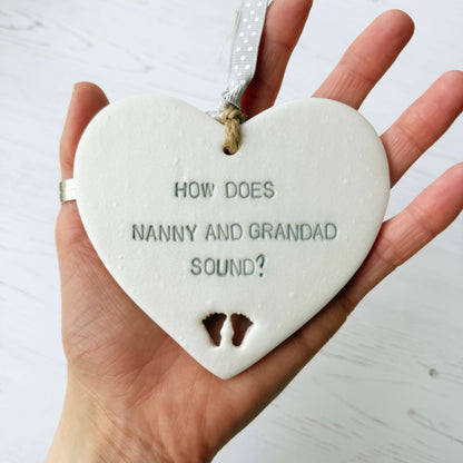 Pearlised white clay hanging heart with baby feet cut out of the bottom and grey personalisation, the heart is personalised with HOW DOES NANNY AND GRANDAD SOUND?