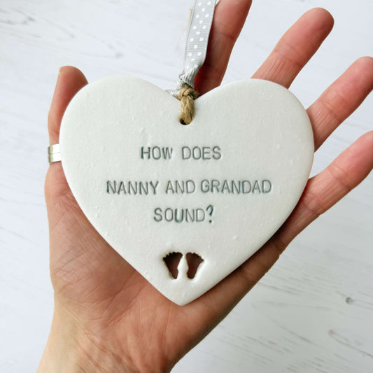Pearlised white clay hanging heart with baby feet cut out of the bottom and grey personalisation, the heart is personalised with HOW DOES NANNY AND GRANDAD SOUND?