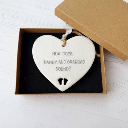 Pearlised white clay hanging heart with baby feet cut out of the bottom and grey personalisation, the heart is personalised with HOW DOES NANNY AND GRANDAD SOUND?