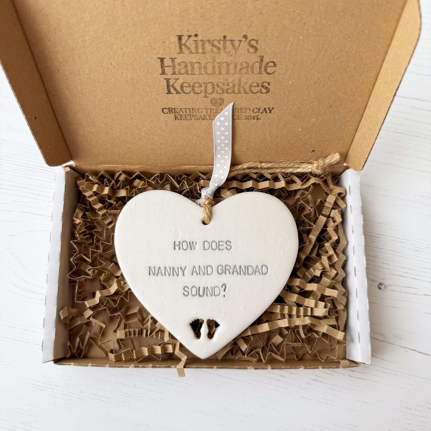Pearlised white clay hanging heart with baby feet cut out of the bottom and grey personalisation, the heart is personalised with HOW DOES NANNY AND GRANDAD SOUND?