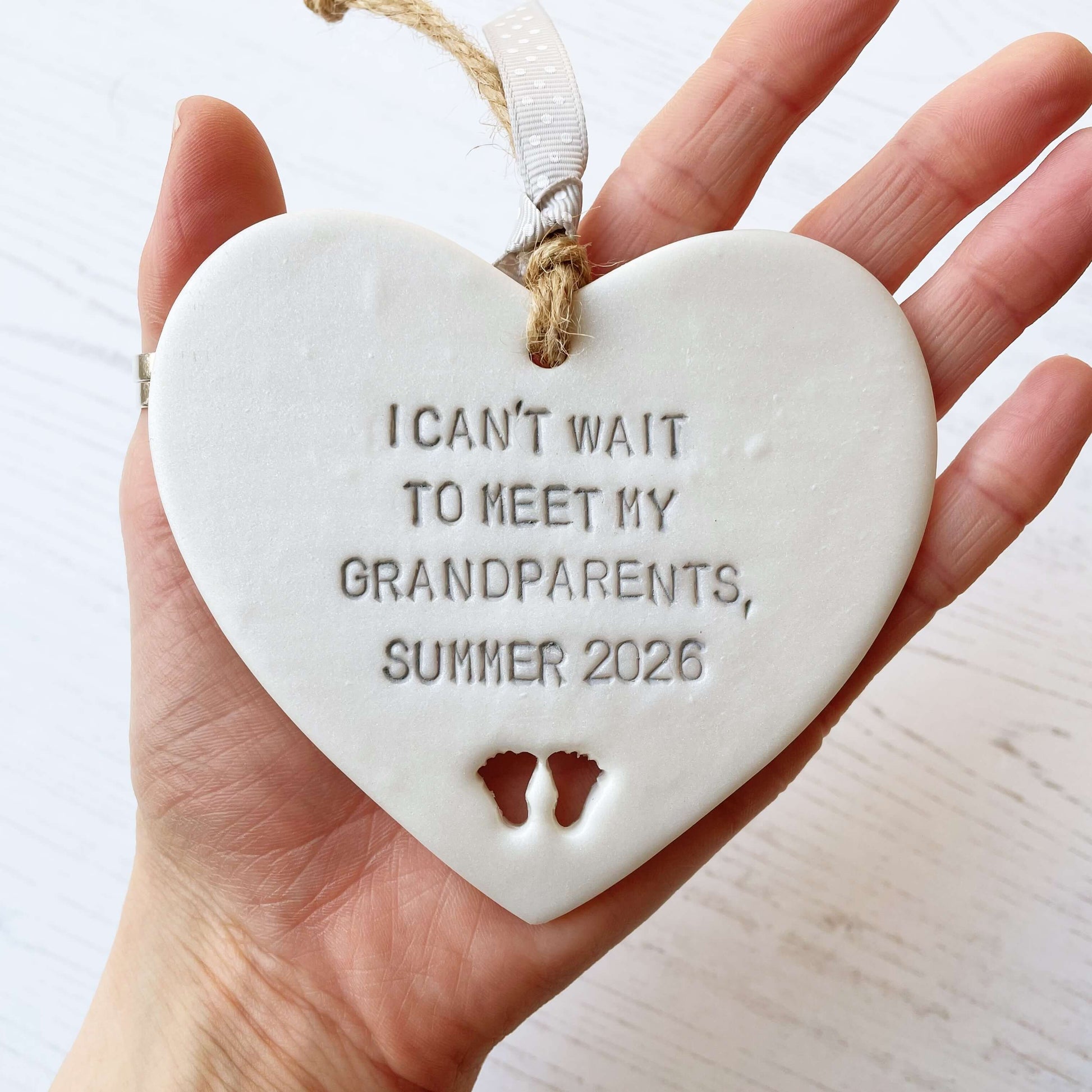 Pearlised white clay hanging heart with baby feet cut out of the bottom and grey personalisation, the heart is personalised with I CAN’T WAIT TO MEET MY GRANDPARENTS, SUMMER 2026