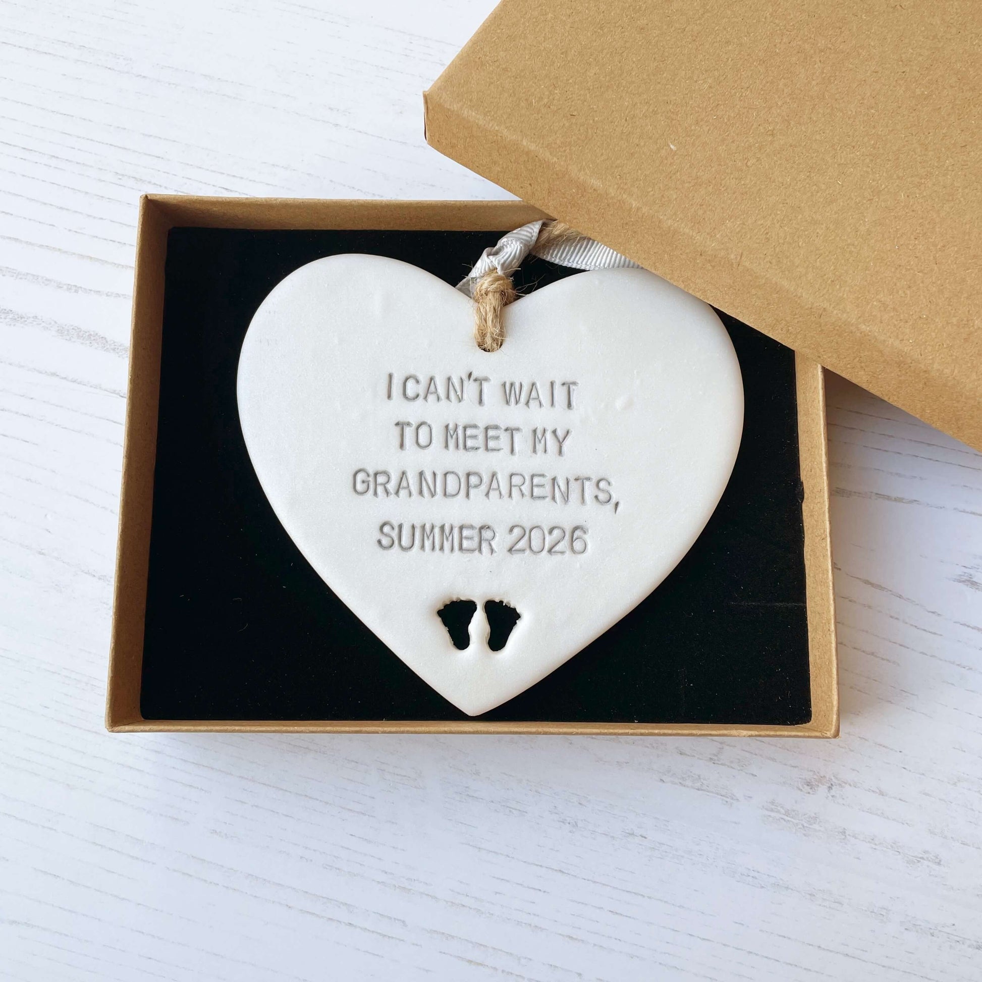 Pearlised white clay hanging heart with baby feet cut out of the bottom and grey personalisation, the heart is personalised with I CAN’T WAIT TO MEET MY GRANDPARENTS, SUMMER 2026
