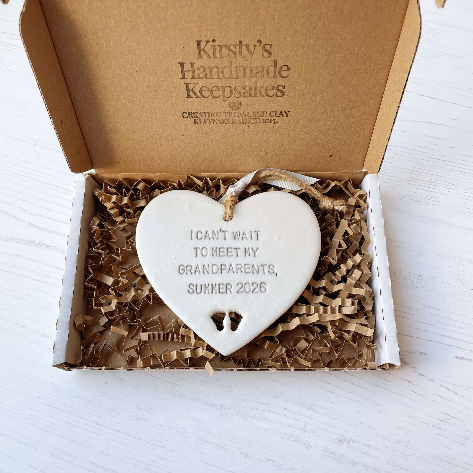 Pearlised white clay hanging heart with baby feet cut out of the bottom and grey personalisation, the heart is personalised with I CAN’T WAIT TO MEET MY GRANDPARENTS, SUMMER 2026