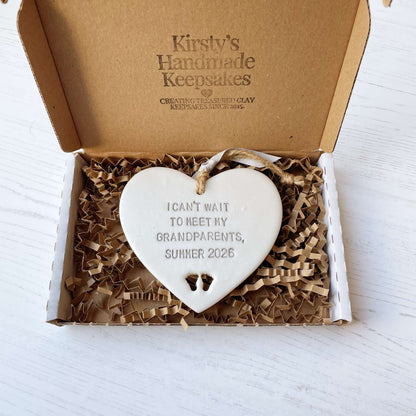 Pearlised white clay hanging heart with baby feet cut out of the bottom and grey personalisation, the heart is personalised with I CAN’T WAIT TO MEET MY GRANDPARENTS, SUMMER 2026