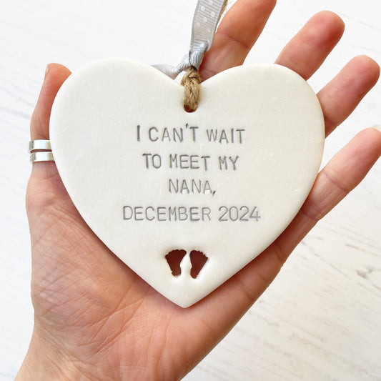 Pearlised white clay hanging heart with baby feet cut out of the bottom and grey personalisation, the heart is personalised with I CAN’T WAIT TO MEET MY NANA, DECEMBER 2024