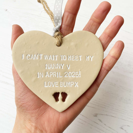 Beige clay hanging heart with baby feet cut out of the bottom and white personalisation, the heart is personalised with I CAN’T WAIT TO MEET MY NANNY V IN APRIL 2025! LOVE BUMP X