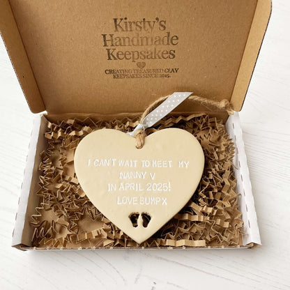 Beige clay hanging heart with baby feet cut out of the bottom and white personalisation, the heart is personalised with I CAN’T WAIT TO MEET MY NANNY V IN APRIL 2025! LOVE BUMP X