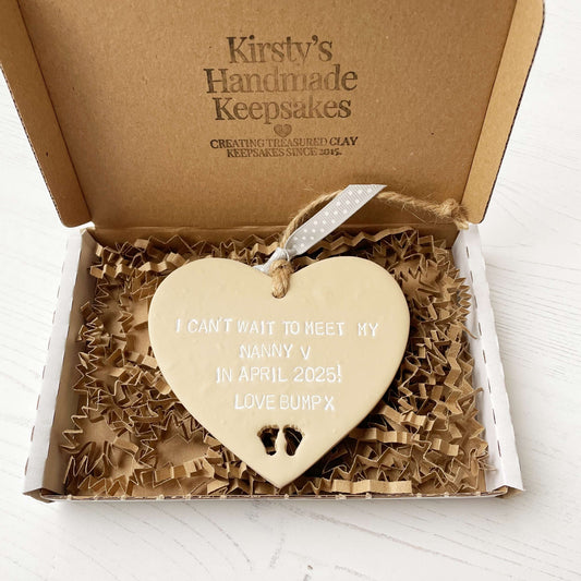 Beige clay hanging heart with baby feet cut out of the bottom and white personalisation, the heart is personalised with I CAN’T WAIT TO MEET MY NANNY V IN APRIL 2025! LOVE BUMP X