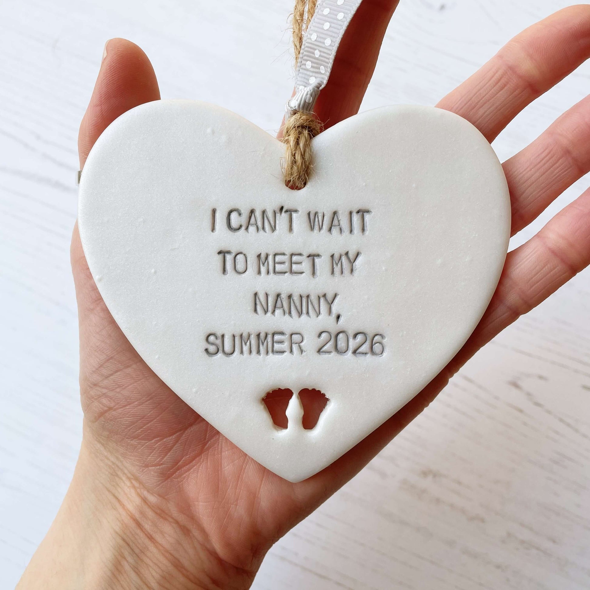 Pearlised white clay hanging heart with baby feet cut out of the bottom and grey personalisation, the heart is personalised with I CAN’T WAIT TO MEET MY NANNY, SUMMER 2026