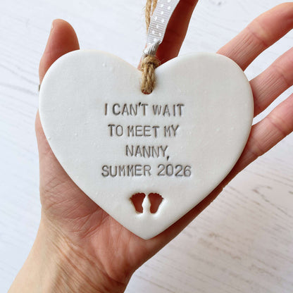 Pearlised white clay hanging heart with baby feet cut out of the bottom and grey personalisation, the heart is personalised with I CAN’T WAIT TO MEET MY NANNY, SUMMER 2026