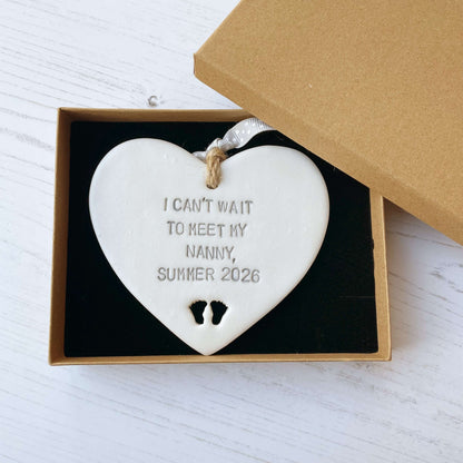 Pearlised white clay hanging heart with baby feet cut out of the bottom and grey personalisation, the heart is personalised with I CAN’T WAIT TO MEET MY NANNY, SUMMER 2026