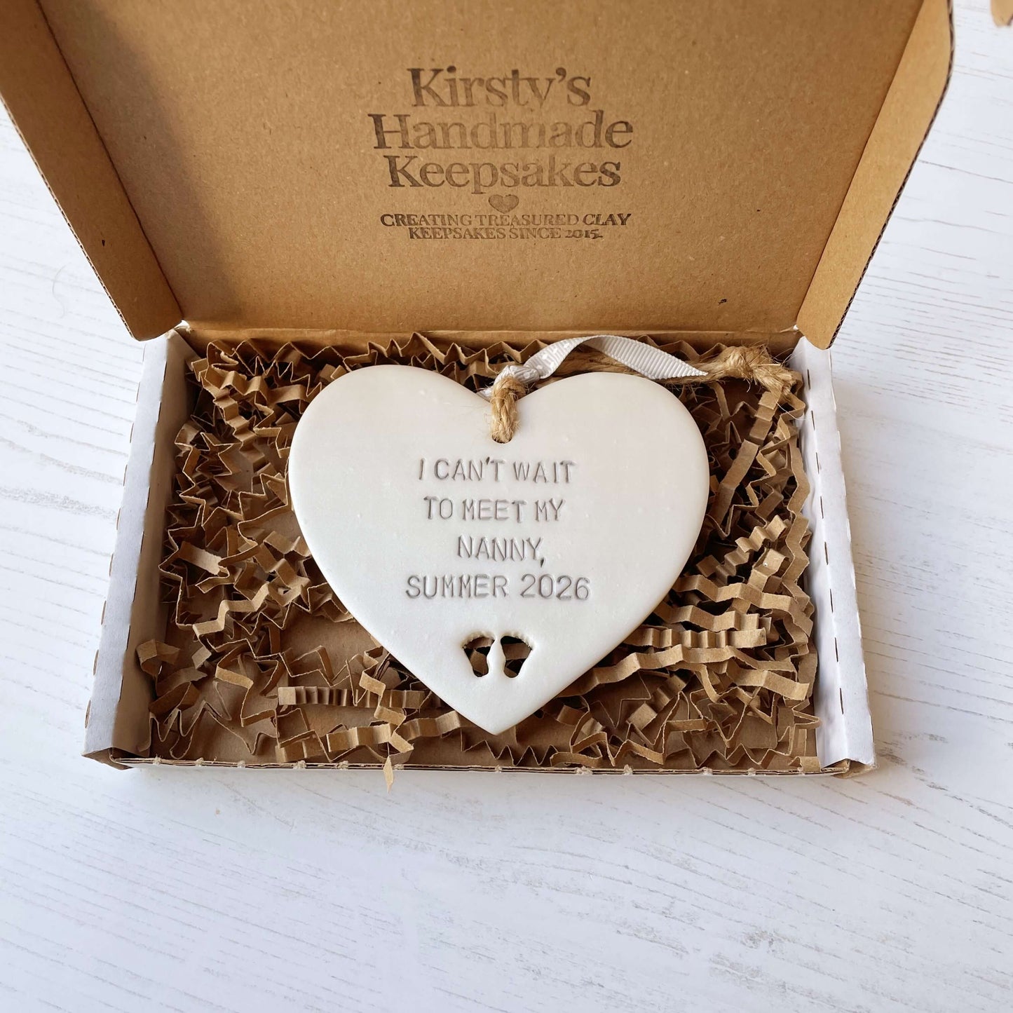 Pearlised white clay hanging heart with baby feet cut out of the bottom and grey personalisation, the heart is personalised with I CAN’T WAIT TO MEET MY NANNY, SUMMER 2026