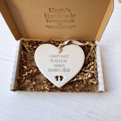 Pearlised white clay hanging heart with baby feet cut out of the bottom and grey personalisation, the heart is personalised with I CAN’T WAIT TO MEET MY NANNY, SUMMER 2026