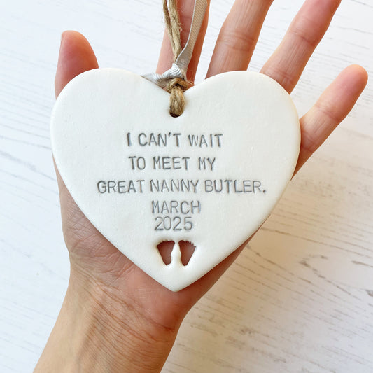 Pearlised white clay hanging heart with baby feet cut out of the bottom and grey personalisation, the heart is personalised with I CAN’T WAIT TO MEET MY GREAT NANNY BUTLER. MARCH 2025